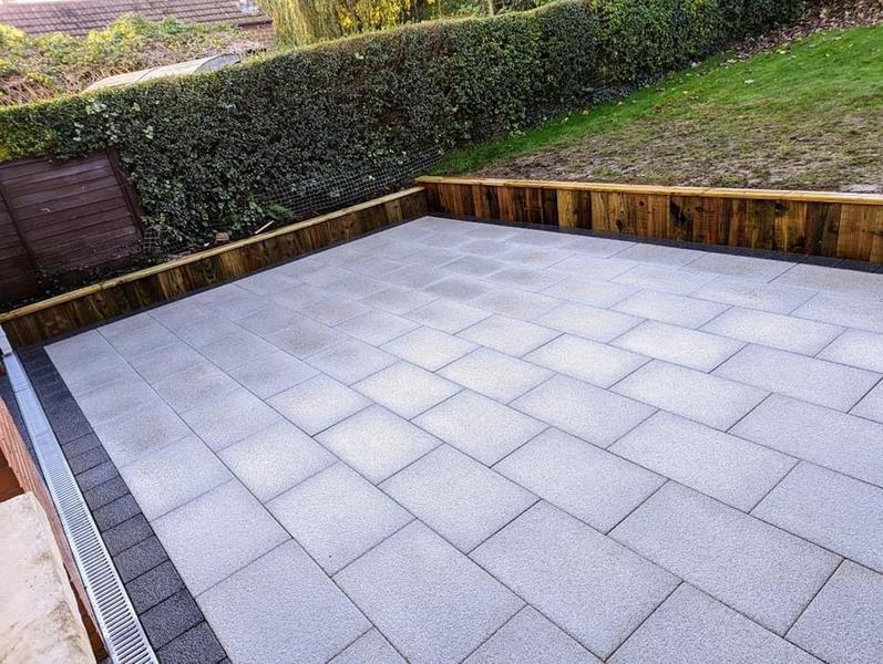 Tobermore Paving Project Large paved garden patio with light grey rectangular slabs, timber retaining border and hedges beyond